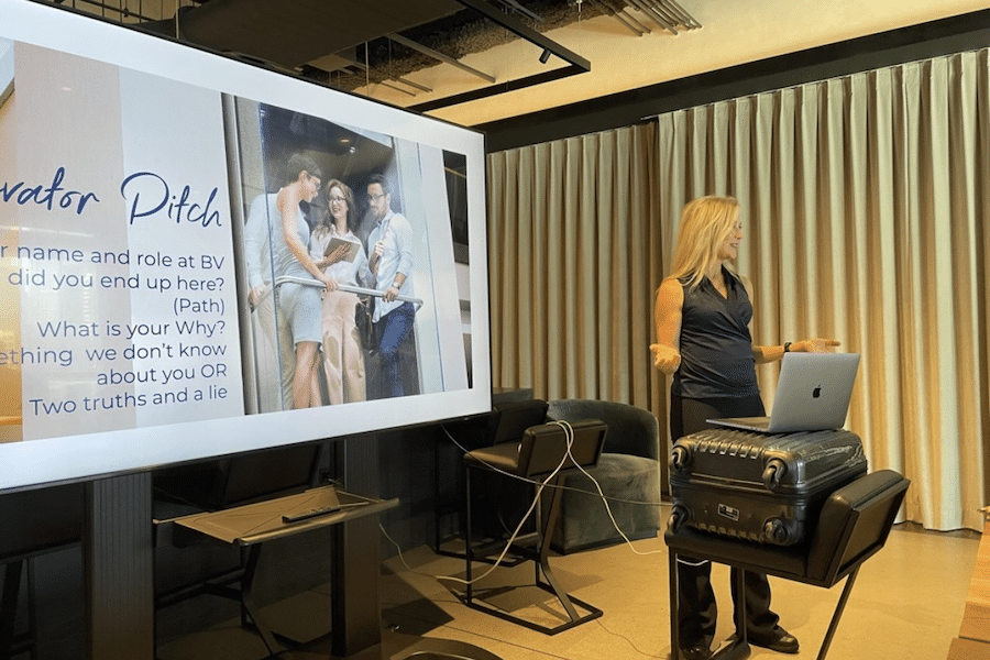 Blonde, white-passing woman teaching with a slideshow that reads "elevator pitch" behind her.