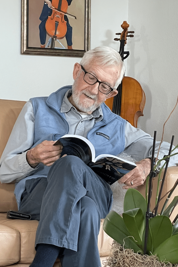 Older, white-passing man with white hair and black-framed glasses, sitting on a couch reading The Voice Coach's Toolkit.